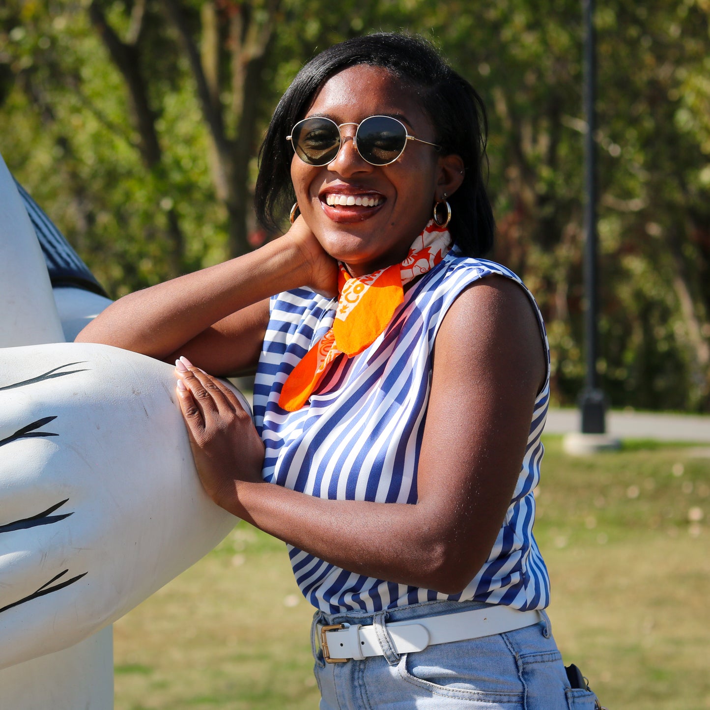 Woman wearing sunglasses and a striped shirt with an orange scarf, standing outdoors.