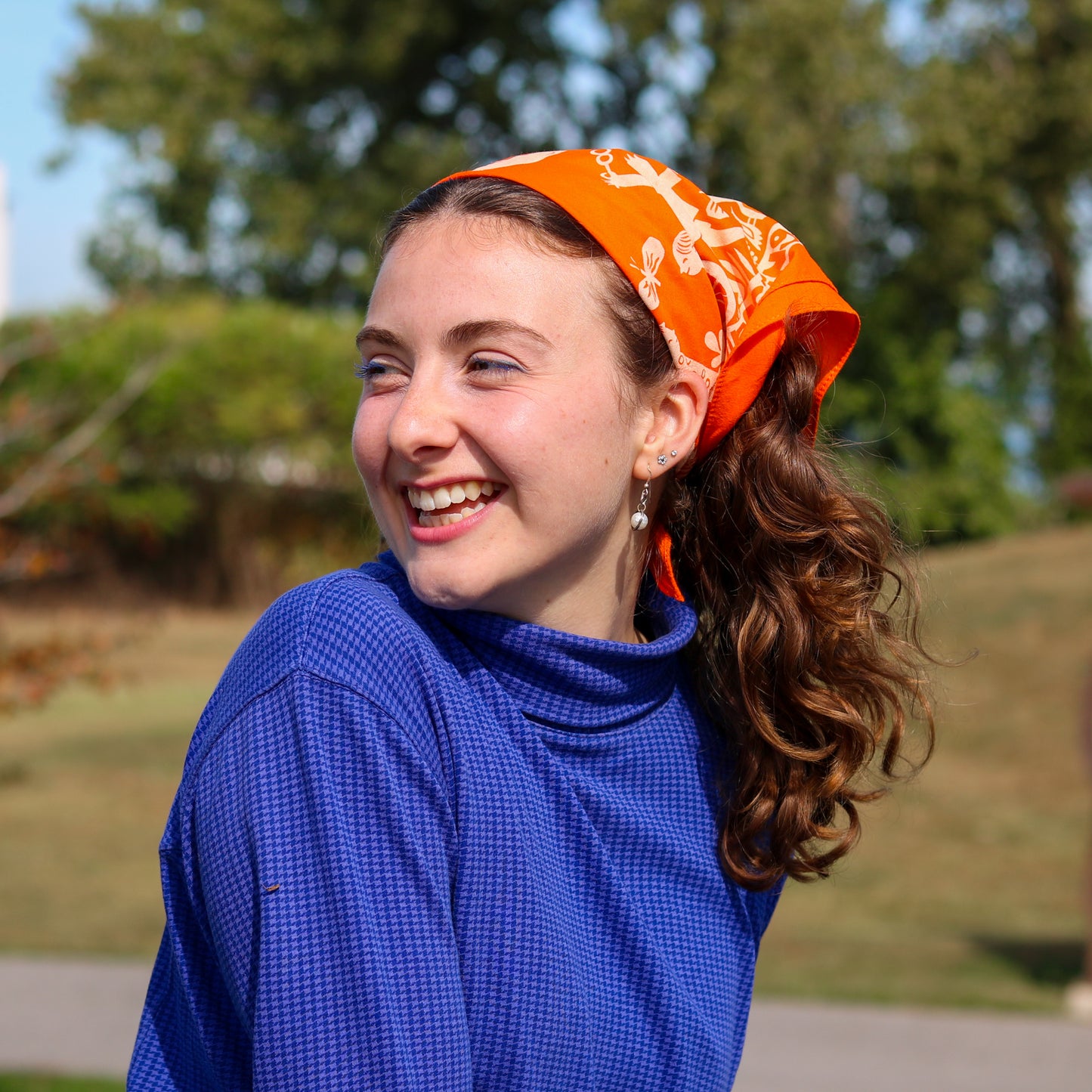 Woman wearing an orange headscarf with a pattern, smiling outdoors.